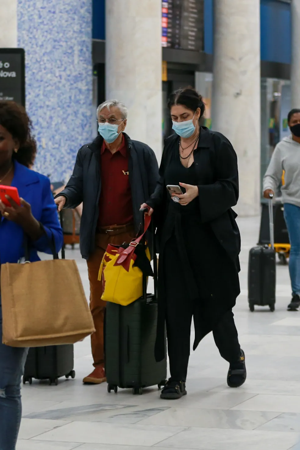 Caetano Veloso aparece com malas nas mãos e máscara de proteção no rosto. Paula Lavigne segura um celular com uma das mãos, enquanto leva as malas na outra