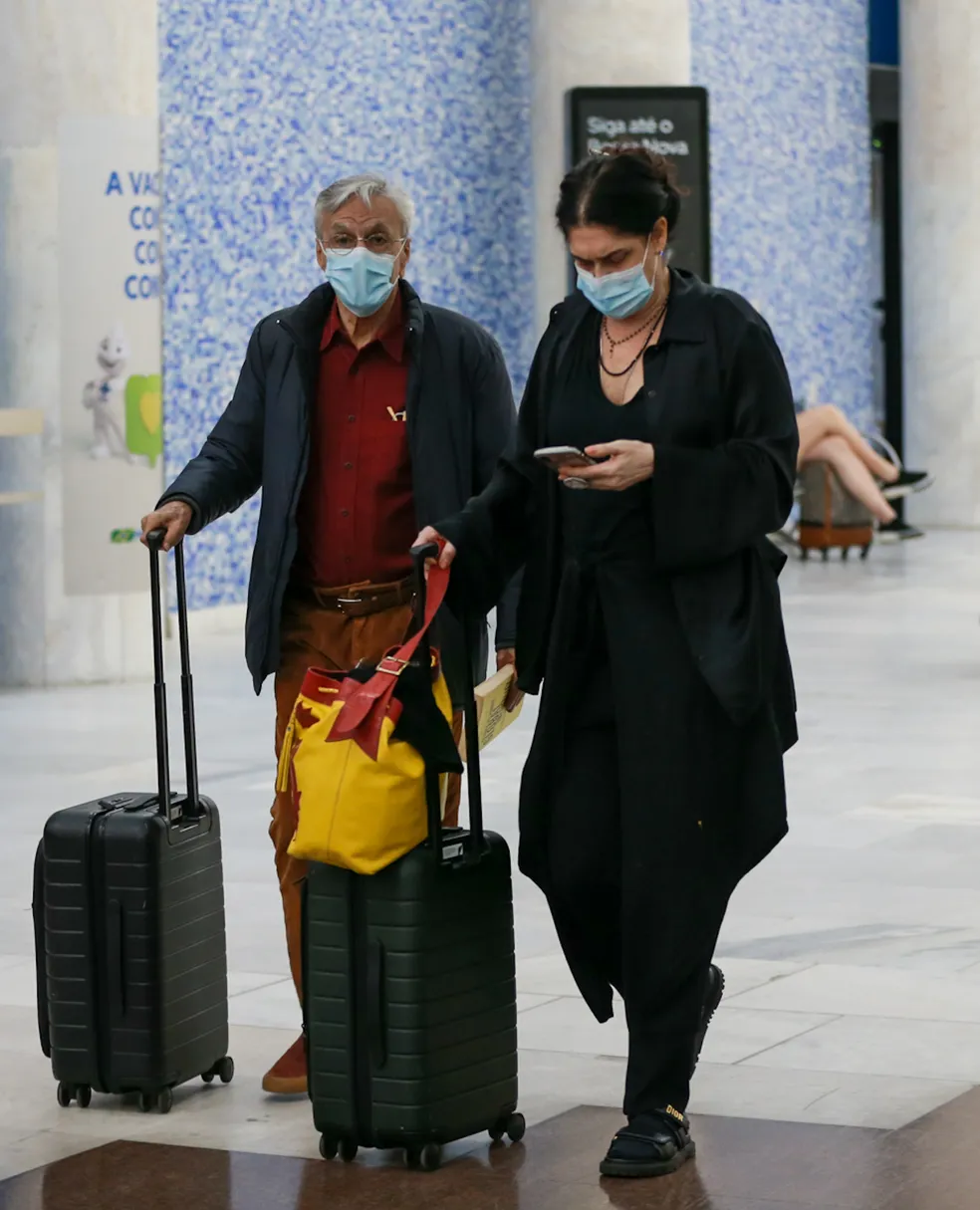 Caetano Veloso aparece com malas nas mãos e máscara de proteção no rosto. Paula Lavigne segura um celular com uma das mãos, enquanto leva as malas na outra