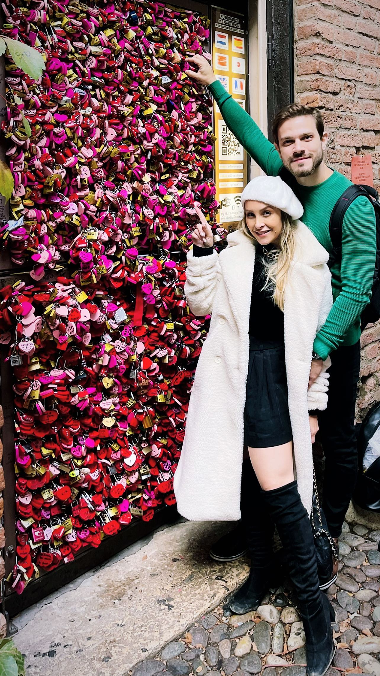 Carla Diaz e Felipe Becari posam ao lado de uma parede cheia de cadeados rosas