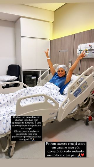Foto de Mileide Mihaile na cama do hospital. Ela aparece com bata hospitalar e um touca branca na cabeça. Deitada na cama, ela coloca as mãos para cima, em sinal de celebração pelo sucesso da cirurgia