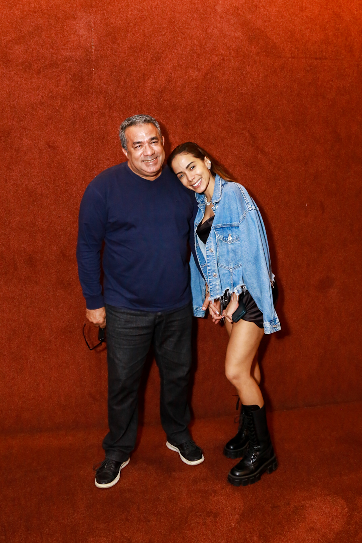 Foto de Anitta com o pai, Mauro Machado, em frente a um tecido vermelho. Ela usa uma jaqueta jeans sobre um vestido preto, sem maquiagem, com os cabelos penteados para trás. Ele veste um suéter azul e calça jeans.