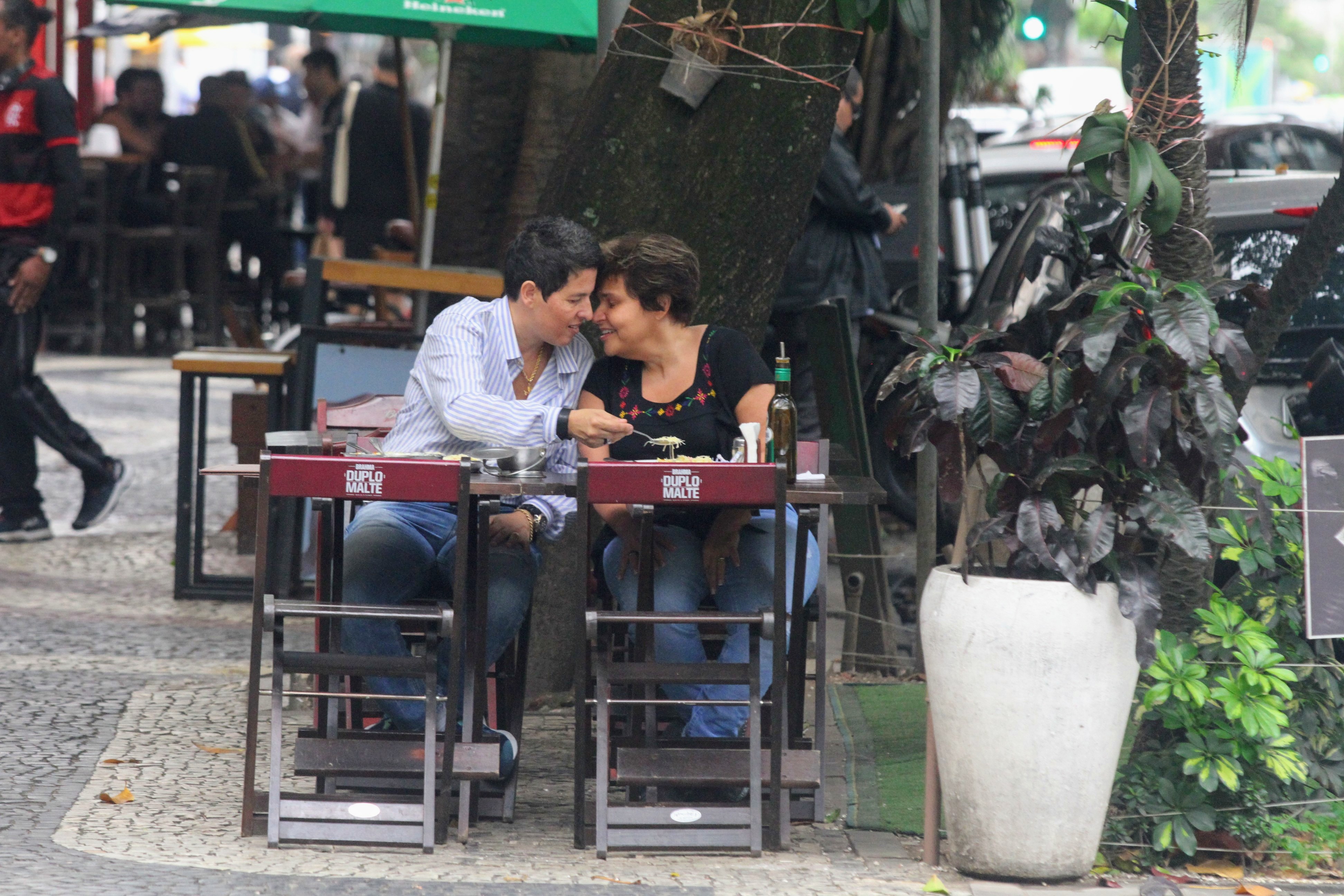 Foto de Adriane Bonato de braço dado com Claudia Rodrigues sentadas em uma mesa de bar trocando carinhos enquanto almoçam