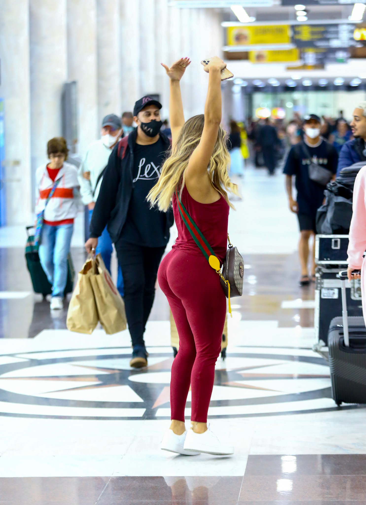 Foto de Lexa no aeroporto com um conjunto de legging e top cor de vinho, usando máscara e fazendo um passo de dança