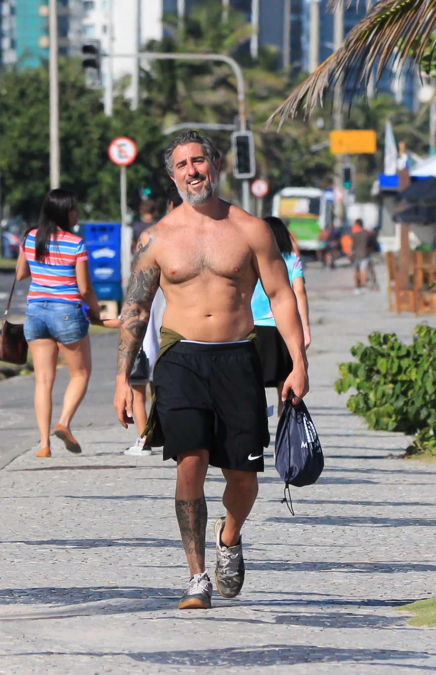 Foto de Marcos Mion andando sem camisa à beira da praia com corpo musculoso à mostra e sorridente