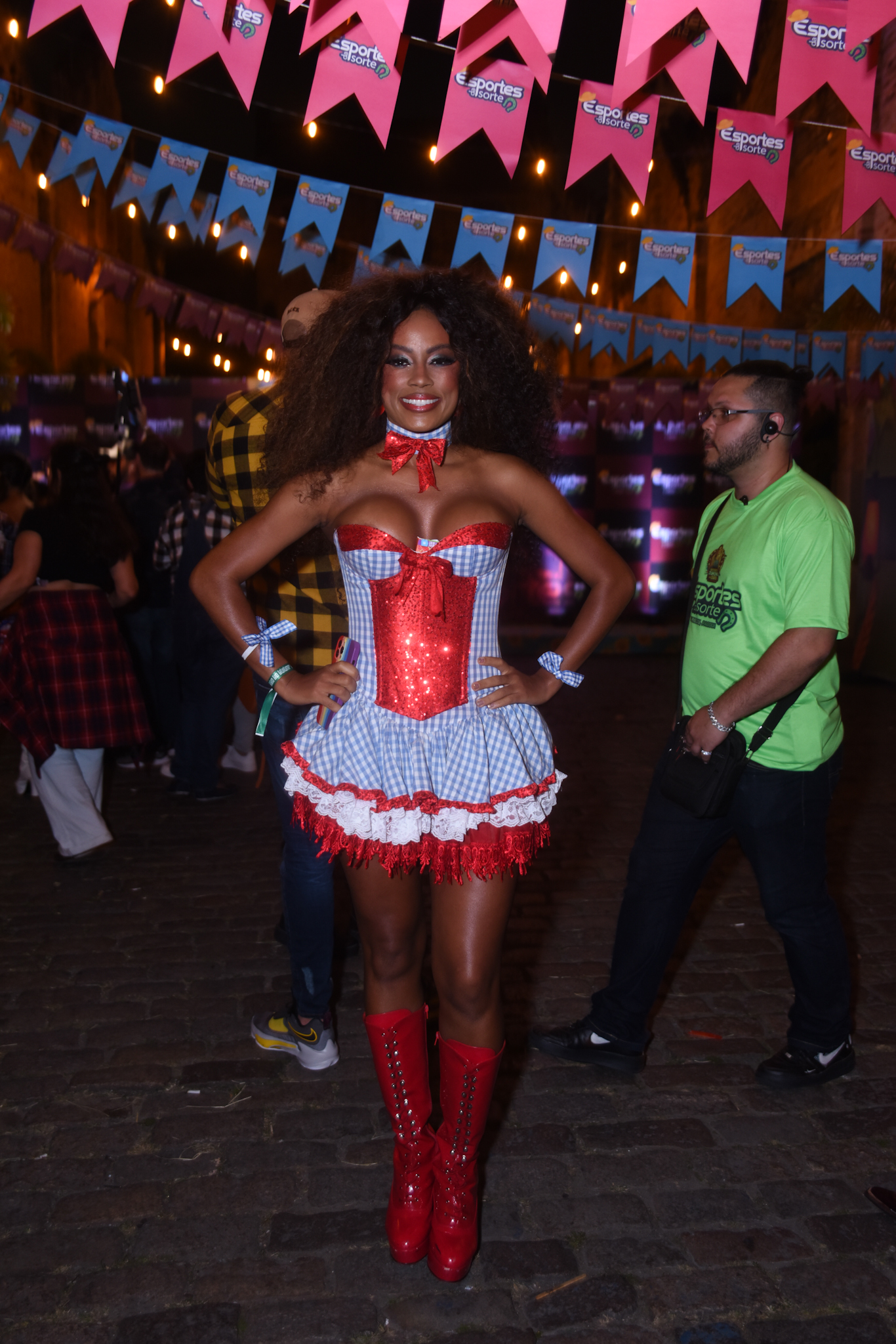 Foto da ex-BBB Lumena Aleluia de vestido xadrez azul com detalhes em vermelho, bota vermelha, cabelos soltos e volumosos, segurando celular com cores do arco-íris, com bandeiras de festa junina ao fundo
