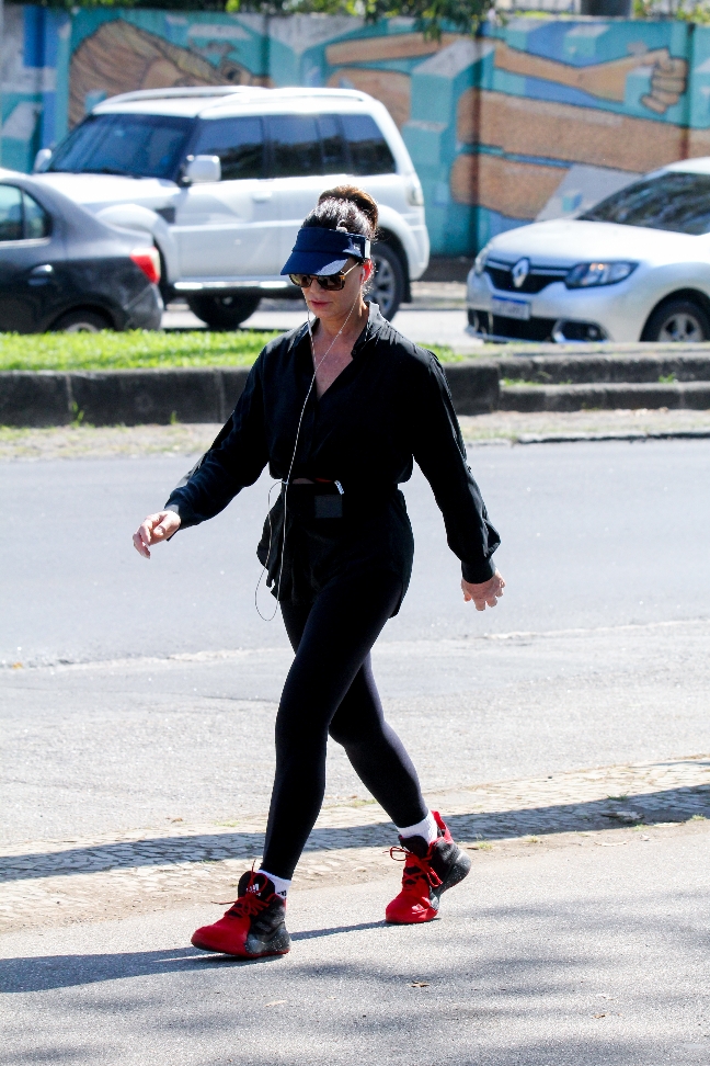 Foto de Luiza Brunet caminhando com uma blusa preta, calça legging preta, tênis vermelho, viseira azul e óculos escuros