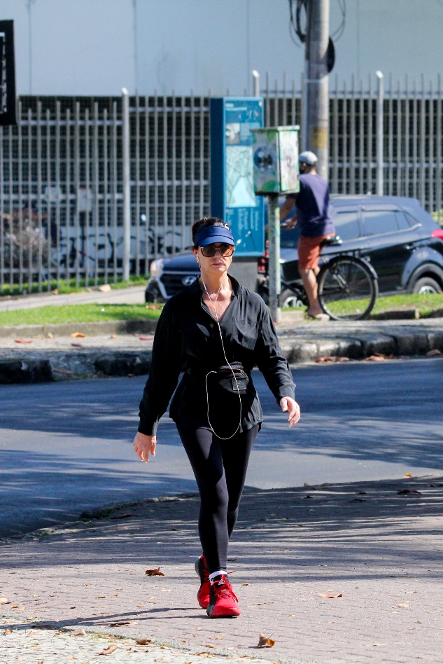 Foto de Luiza Brunet caminhando com uma blusa preta, calça legging preta, tênis vermelho, viseira azul e óculos escuros