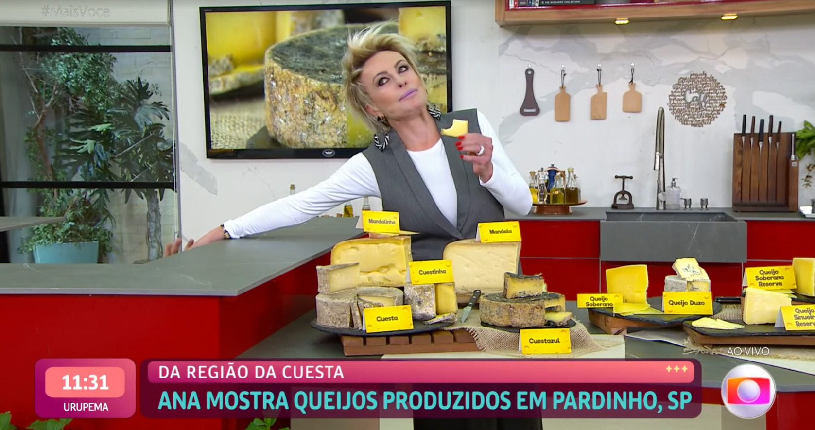 Foto retirada do Globoplay de Ana Maria Braga no estúdio do 'Mais Você' esticando a mão para entregar um pedaço de queijo para o Louro Mané, cuja mão aparece