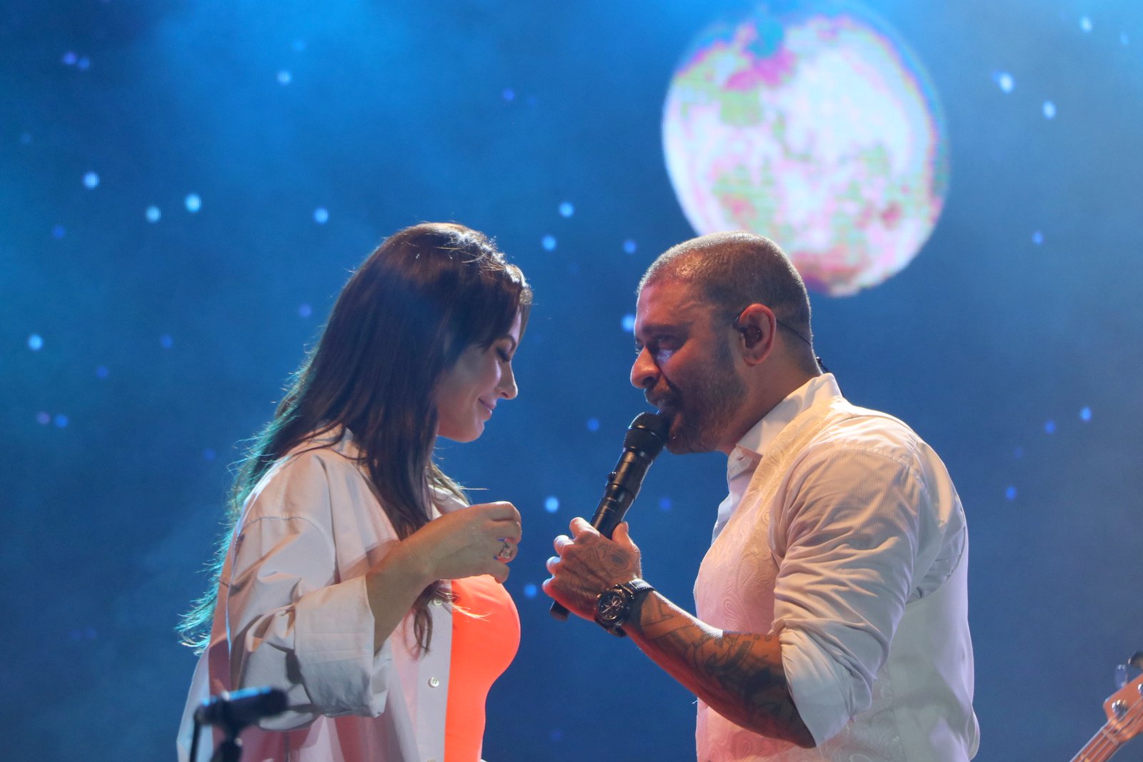 Foto de Paolla Oliveira e Diogo Nogueira no palco de um show, com um telão exibindo uma lua em um céu estrelado ao fundo enquanto o cantor canta no microfone e ela dança