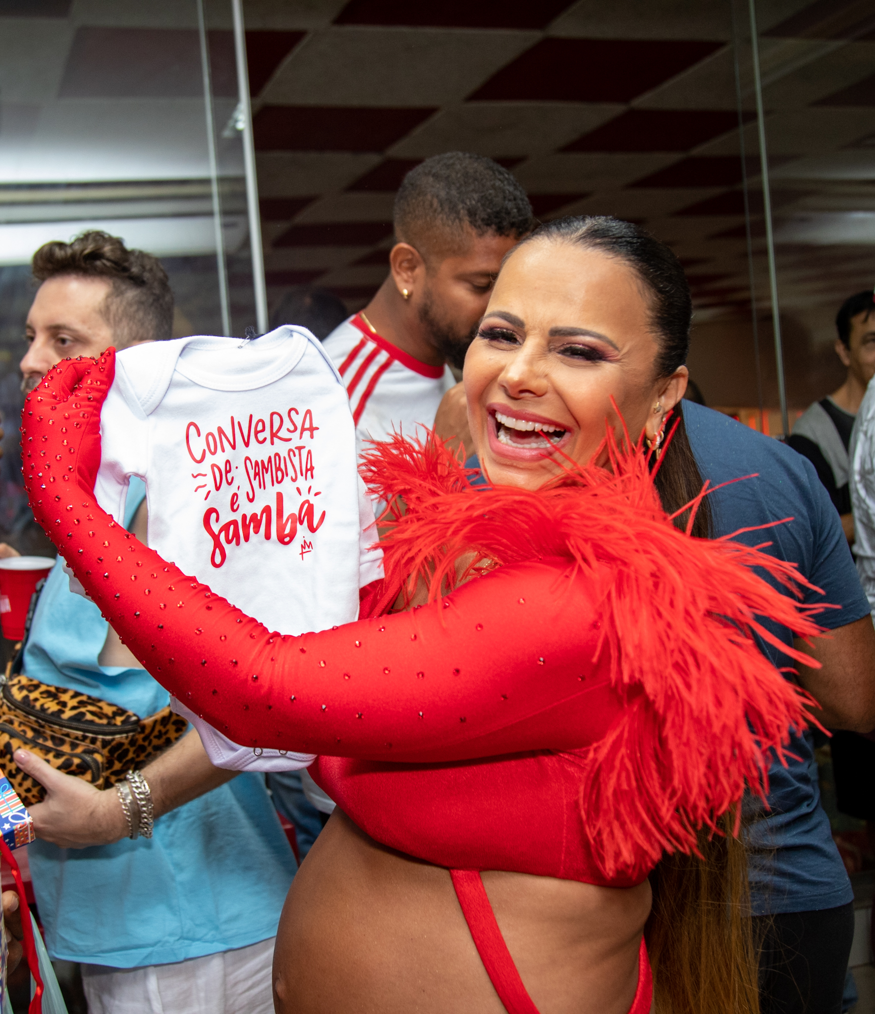 Foto de Viviane Araújo com conjunto de top e calça vermelhos e a barriga de grávida à mostra segurando um macacãozinho para o bebê com os dizeres: "Conversa de sambista é samba"